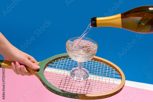 Photography Champagne pouring into coupe glass on vintage tennis racket over pastel pink and blue background