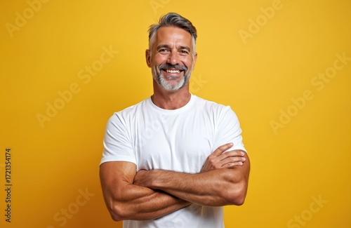 Athletic middle-aged man with grey hair smiles confidently, arms folded, wearing white t-shirt. Muscular build, mature, masculine presence. Positive expression on yellow background. Health, fitness,