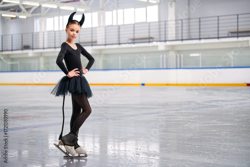 Young figure skater in a devil costume with a broomstick performing on the ice rink during Halloween