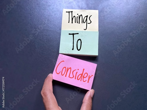 A hand is holding a pink sticky note with the word Consider, which is placed below two other sticky notes with the words Things To. Refers to think about important points before making a decision.