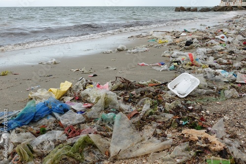 A wide-angle view of a dirty beach with a vast amount of plastic waste, including bottles, bags, and other debris, piled up on the sand near the water's edge.