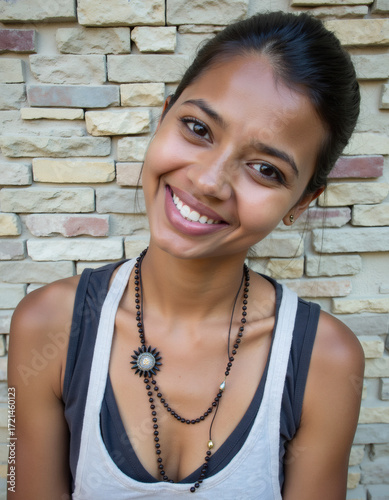 Joyful Young Woman with a Radiant Smile and Unique Beaded Necklace, Tilting Her Head Playfully Against a Rustic Stone Wall