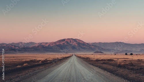 A long, straight desert road vanishes towards a large, imposing mountain range under a soft, pastel sunset sky