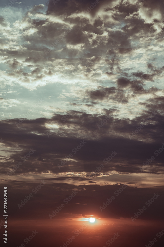 Naklejka premium A vibrant sunset is captured as the sun dips below a dramatic sky filled with dark and light grey clouds. The clouds showcase a mix of textures and tones, creating depth and visual interest