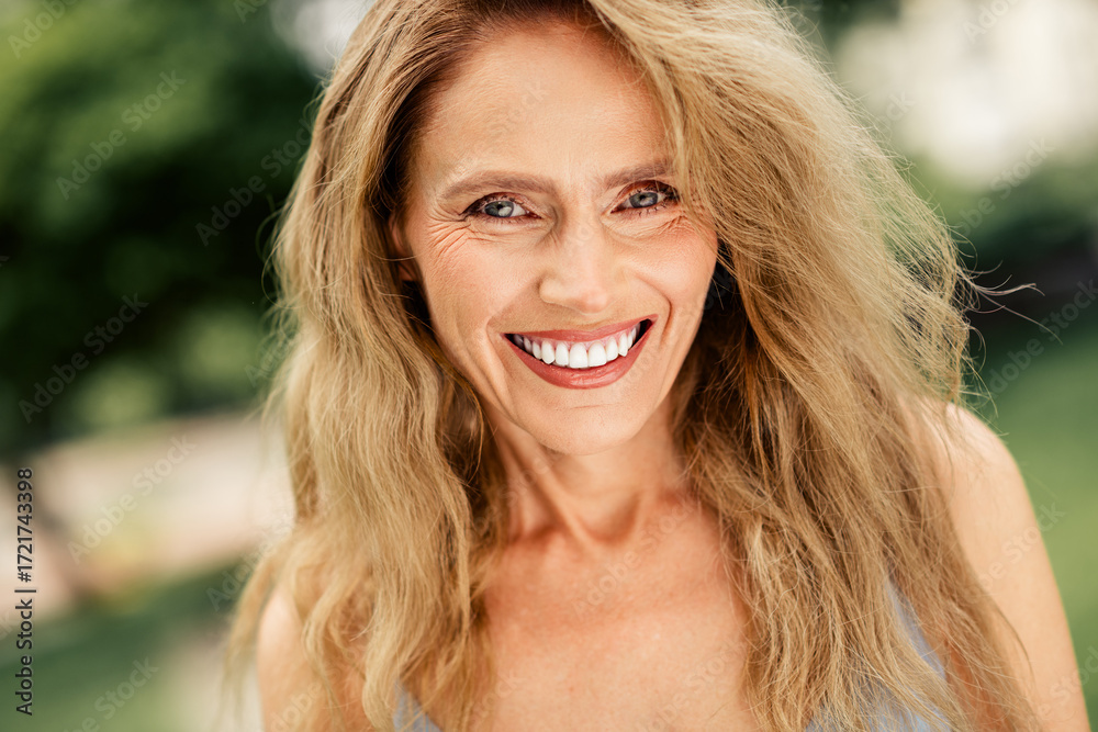 Fototapeta premium Charming middle-aged woman smiling warmly outdoors with natural surroundings on a sunny day, radiating happiness