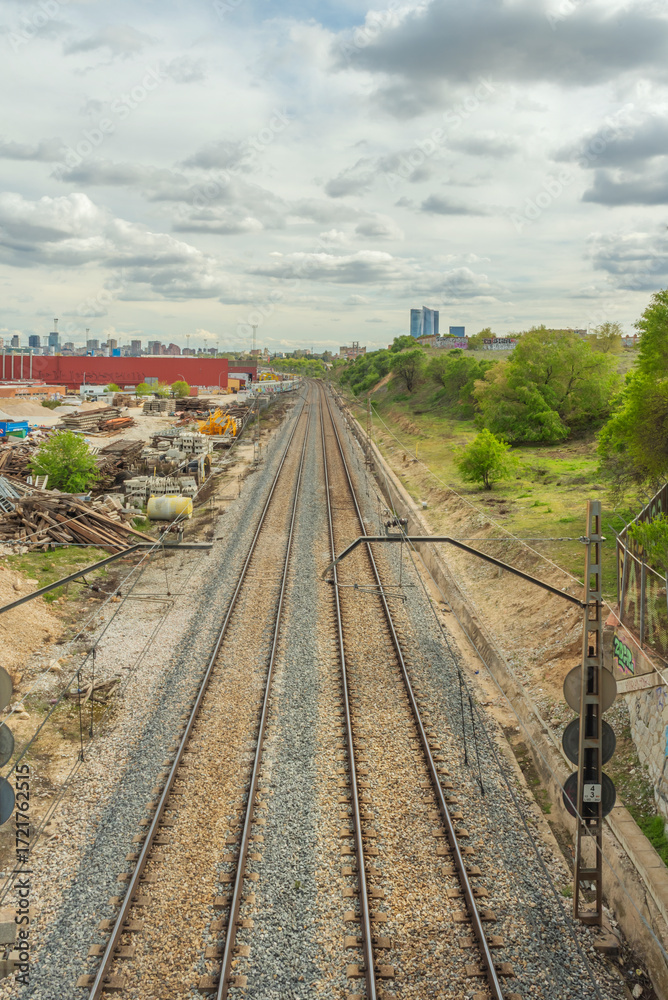 Obraz premium Straight section of commuter rail track with gray ballast and concrete sleepers, flanked by a safety fence and overhead power lines