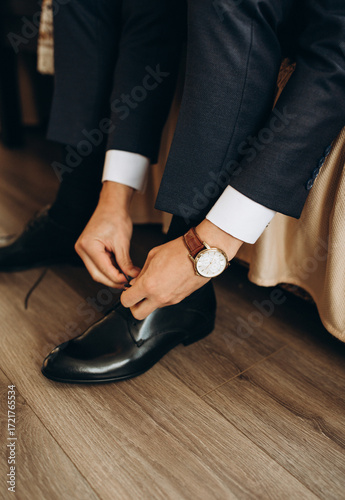 Close-up of groom tying shoelaces on elegant black leather shoes. Man in dark suit with stylish wristwatch preparing for wedding ceremony. Detail shot of men’s fashion and wedding morning routine.