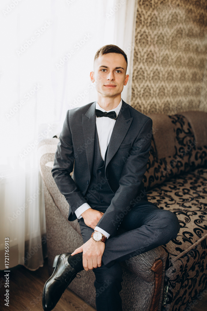 Fototapeta premium Groom in stylish dark suit with bow tie sitting indoors on a sofa. Young man with classic watch and polished shoes, posing confidently on his wedding day. Elegant portrait in formal attire.