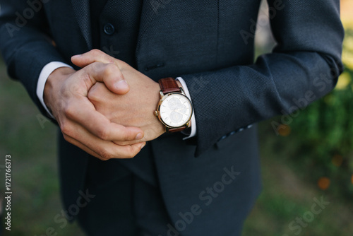Close-up of groom in stylish dark suit with hands clasped, wearing an elegant wristwatch with brown leather strap. Classic men’s fashion detail for wedding, business, or formal occasions.
