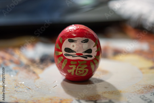 Close-up photograph of a Daruma doll, its red exterior and painted features standing out against a textured, blurred background