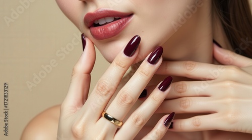 Close-up of a woman's manicured hands with glossy burgundy almond nails, gold ring, and soft beige background, capturing refined elegance and modern beauty in natural light.