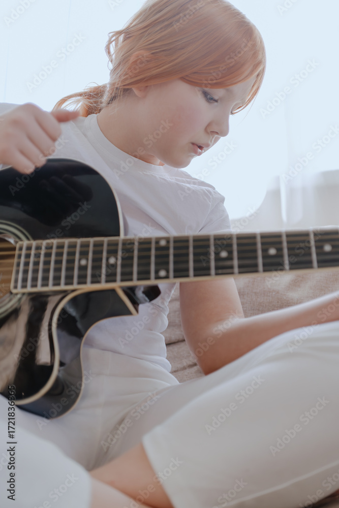 Fototapeta premium A teenage girl plays guitar in a bright living room, captured in an authentic moment of musical passion. Real photography showcasing genuine youth creativity and emotion