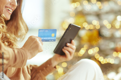 A smiling woman holds a credit card and smartphone, embodying festive holiday online shopping. A blurred Christmas tree with warm bokeh lights creates a cozy atmosphere perfect for e-commerce themes.