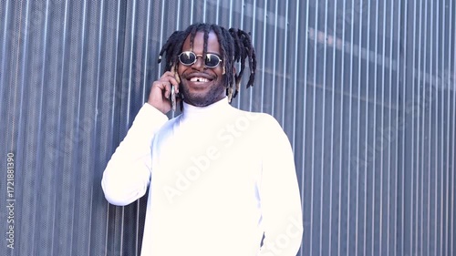 Young black man with dreadlocks and sunglasses smiling while talking on his smartphone
