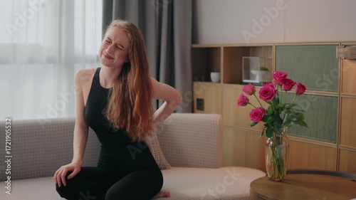 Young woman on a sofa clutches her lower back in pain, painful suffering from injury, backache discomfort, grimacing from a sudden twinge caused by a pinched nerve or muscle strain, lower back pain
