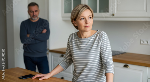 Middle aged woman looking thoughtful while her husband stands in the background arms crossed in their kitchen marital problems relationship issues