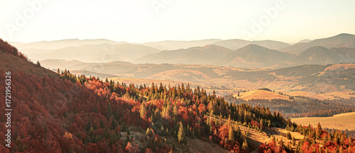 A breathtaking panoramic view of a mountain range in the Carpathian Mountains during a hazy autumn sunset, with vibrant fall foliage on the hillsides illuminated by the warm, golden light.
