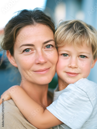 Close-up portrait of a loving middle-aged mother holding her young son cheek to cheek, both smiling and looking at the camera, capturing a warm, affectionate family moment