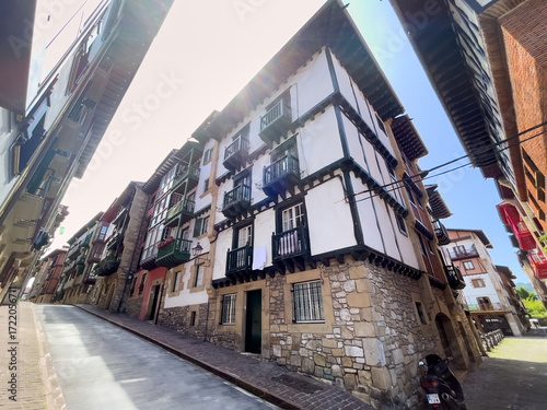 Obraz na płótnie Calle estrecha en pendiente con fachada de entramado de madera y base de piedra, balcones y una motocicleta en un entorno histórico iluminado por luz suave