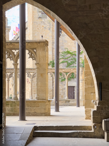 Fotografía Arco apuntado que enmarca patio con arcadas góticas y columnas esbeltas, juego de luz y sombra sobre la piedra