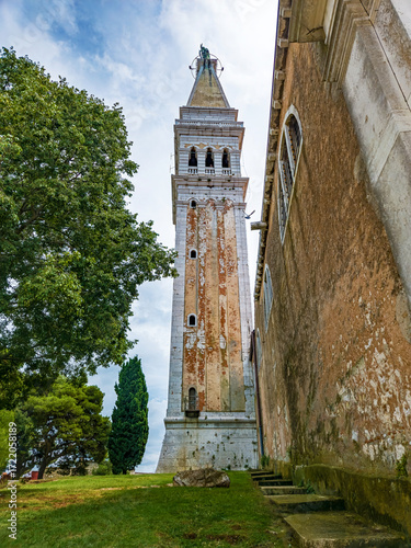 Historic bell tower in Rovinj, Croatia rises above lush greenery and weathered stone walls, framed by dramatic clouds and Mediterranean charm.