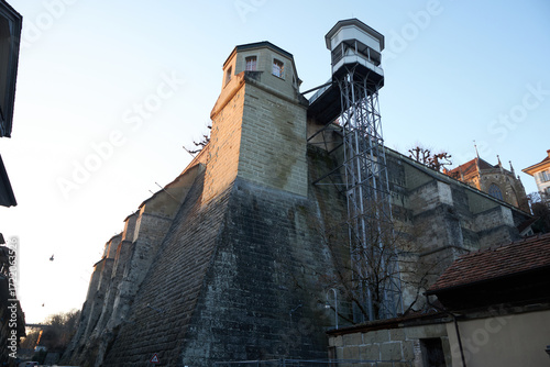 Φωτογραφία Fortaleza de piedra de muros inclinados y torres altas, combinada con un ascensor metálico moderno que asciende por la fachada bajo una luz suave