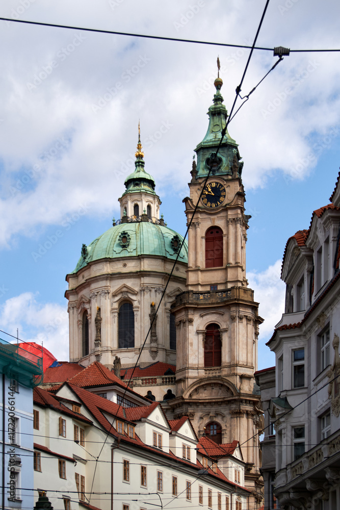 Fototapeta premium Iglesia barroca con torres de cúpulas verdes y reloj dorado, rodeada de edificios históricos de tejados rojos y cables de tranvía bajo un cielo parcialmente nublado.