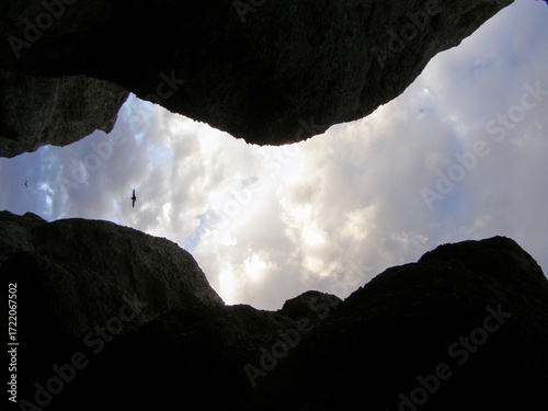 Schilderij op canvas Vista ascendente desde una grieta rocosa hacia un cielo parcialmente nublado, con un ave volando en la distancia enmarcada por las paredes oscuras de la roca