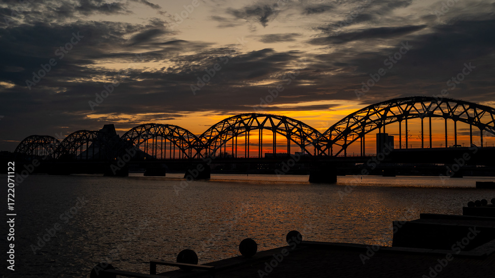 Obraz premium Extremely beautiful and peaceful scene of the Iron Bridge (Dzelzceļa tilts) over the Daugava River at sunset, with its round iron arches standing out against the vibrant orange and blue evening sky.