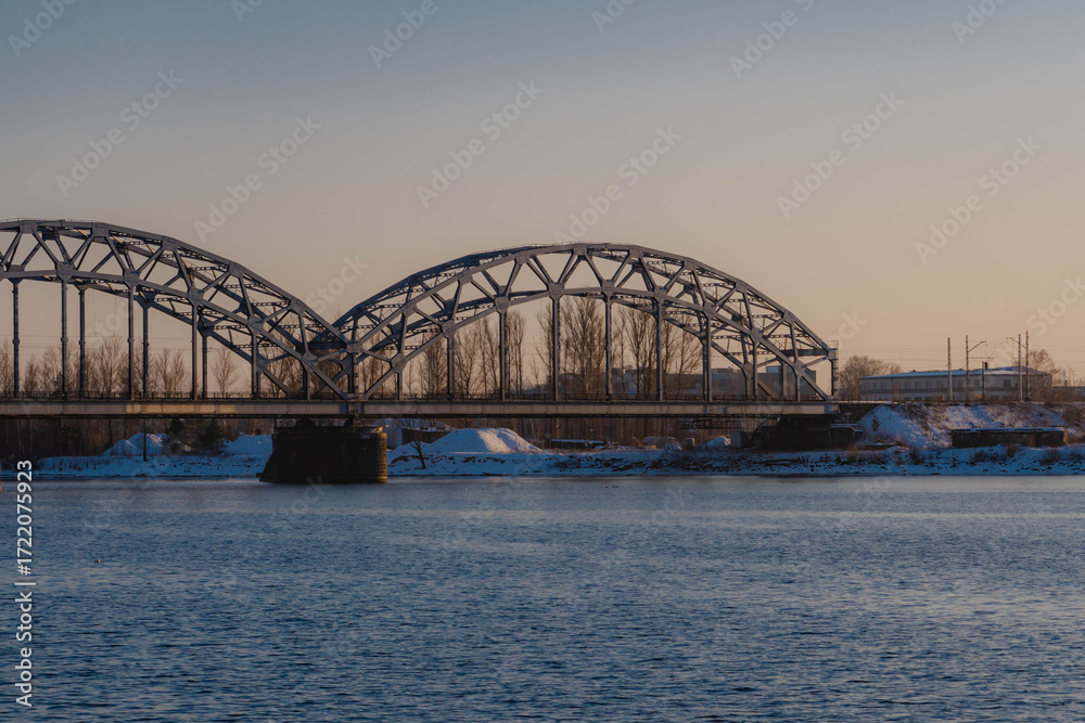 Fototapeta premium Sunset landscape of an iron-framed arched bridge crossing a river in a snowy winter