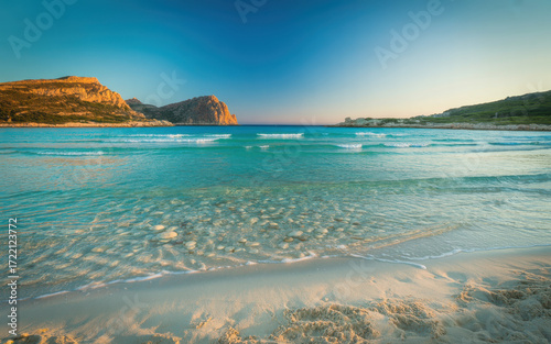 Scenic tropical beach with clear turquoise water gentle waves and rocky cliffs under bright blue sky for travel destination and summer vacation background