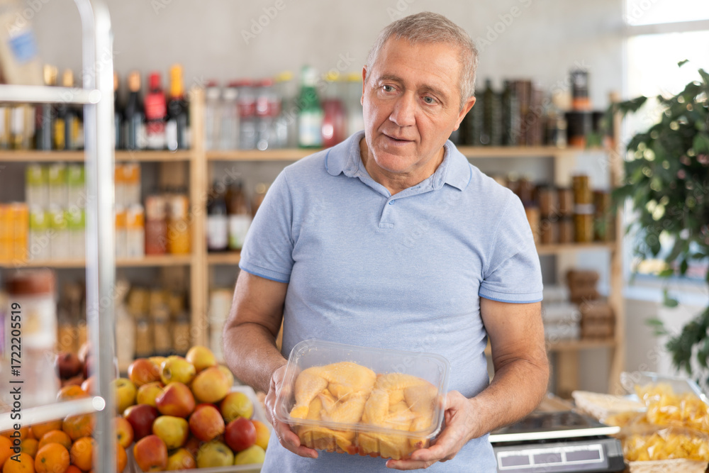 Fototapeta premium Senior man holding plastic containers with poultry half chicken carcass, thinking about buying. He choose ingredients for cooking dinner, thinking before buying, hesitating about choice chicken meat
