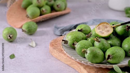 A collection of freshly picked feijoa fruits, some with glistening water droplets, are arranged on a textured white plate resting on a wooden board.