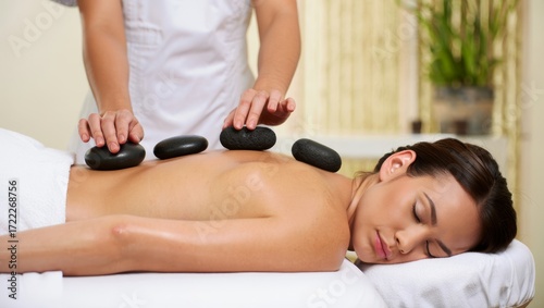 Receiving hot stone massage therapy at a luxury spa, a masseuse placing natural dark basalt stones on a young woman's back for relaxation and wellness treatment