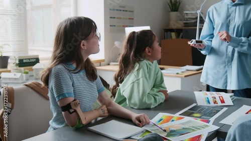 High angle shot of unrecognizable female teacher explaining wind turbine model to attentive students gathered around desk cluttered with drawings and notes in classroom
