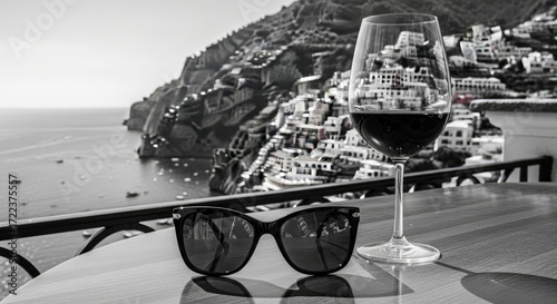 Black and white image of a wine glass filled with red wine and sunglasses on a table overlooking a coastal town and the sea.