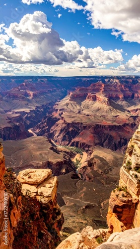 Expansive, sunlit canyon vista showcases layered geological formations under a blue sky with puffy white clouds, extending into the far distance