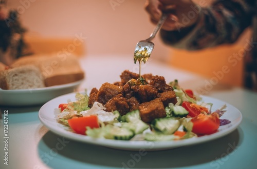 A plate topped with meat and fresh vegetables, a hand using a fork to drizzle dressing. Breads are visible in background, creating soft lighting