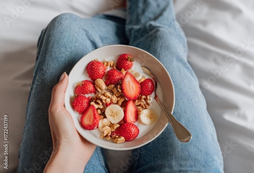 Person holding a bowl of yogurt topped with strawberries, bananas, and walnuts on white bedding