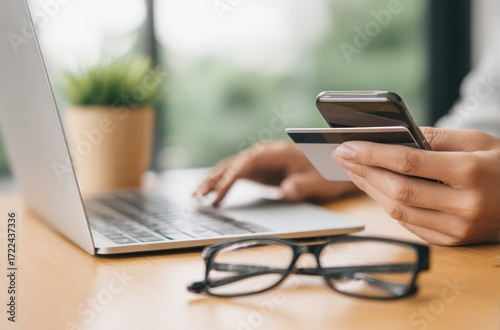 A person is using a smartphone and credit card in front of an open laptop on a wooden table with eyeglasses and a potted plant in the background