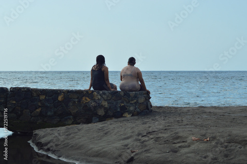 A mother and daughter sit on the sandy bank of a creek with their wet clothes, gazing quietly toward the open ocean horizon under bright daylight.