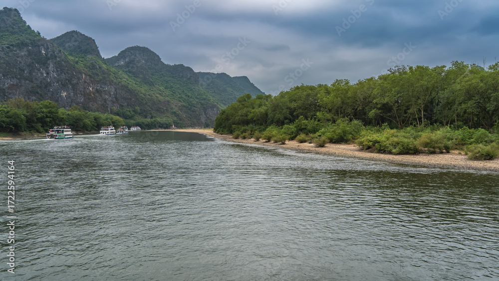 Fototapeta premium Tourist ships sail in the riverbed. Lush green vegetation on the banks of a calm river. Picturesque mountains against the sky and clouds. China. Li River. Li Jiang. Guilin