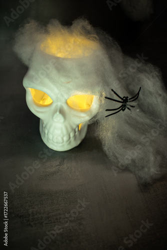 Skull with glowing yellow eyes sits amid cobwebs and a black spider, casting warm light against a dark background. A classic Halloween setup creating a spooky, eerie, and festive atmosphere.