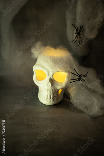 Skull with glowing yellow eyes sits amid cobwebs and a black spider, casting warm light against a dark background. A classic Halloween setup creating a spooky, eerie, and festive atmosphere.