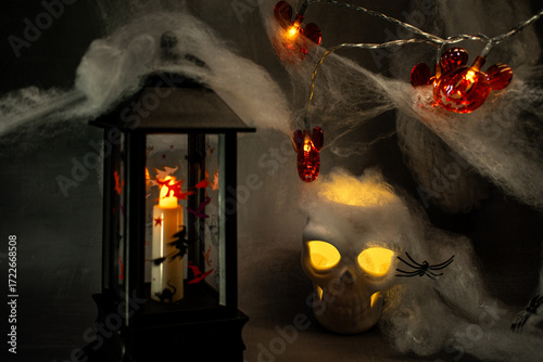 Skull with glowing yellow eyes sits amid cobwebs and a black spider, casting warm light against a dark background. A classic Halloween setup creating a spooky, eerie, and festive atmosphere.