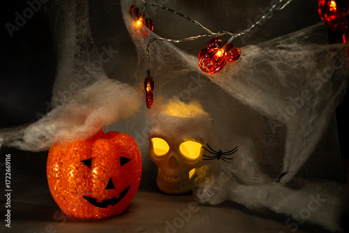 Skull with glowing yellow eyes sits amid cobwebs and a black spider, casting warm light against a dark background. A classic Halloween setup creating a spooky, eerie, and festive atmosphere.