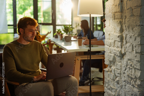 Focused employee working on laptop in modern office with colleagues collaborating