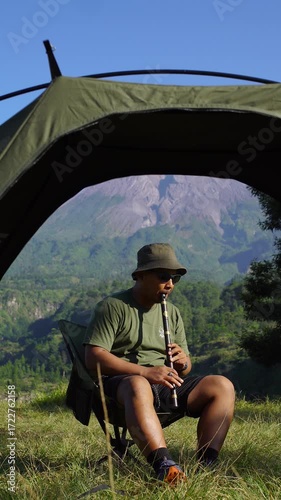 A solo male adventurer plays a traditional instrument called a low whistle against a backdrop of a volcano. A solo camper plays a low whistle on a bright morning against a backdrop of a volcano.