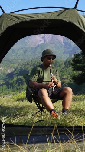 A solo male adventurer plays a traditional instrument called a low whistle against a backdrop of a volcano. A solo camper plays a low whistle on a bright morning against a backdrop of a volcano.