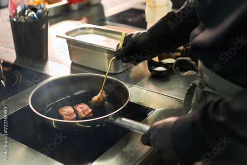 Middle aged Caucasian man wearing black gloves searing pieces of meat in frying pan on stovetop in professional kitchen